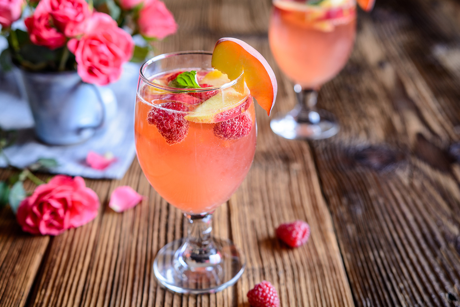 glass of mimosa with raspberries and peaches on wooden table with flower vase