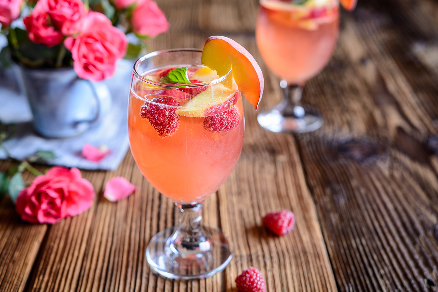 glass of mimosa with raspberries and peaches on wooden table with flower vase 