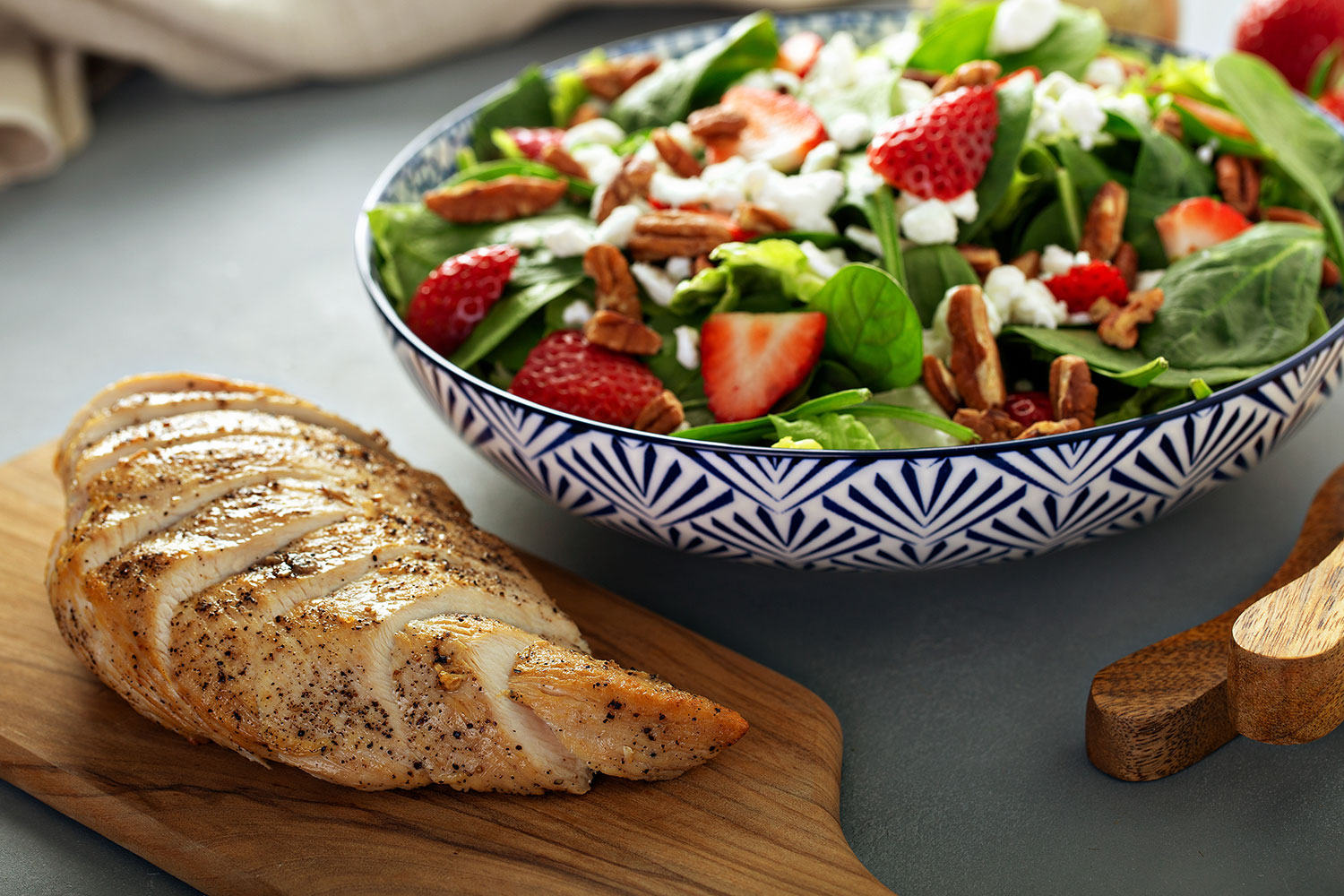 fresh spinach salad with strawberries, feta cheese, halved walnuts in a bowl. slices of grilled chicken displayed on a wood cutting board